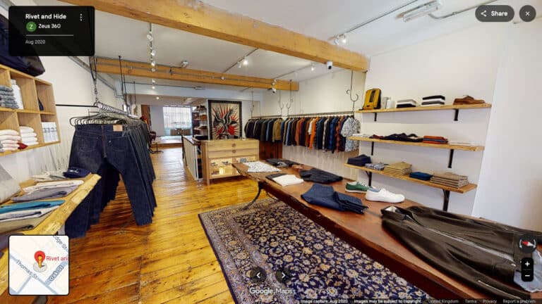 An interactive Google Street View virtual tour through a modern independent boutique shop interior, showcasing Street View navigation arrows on the concrete floor, a woman with curly brown hair browsing a clothing display, and a staff member at a service counter in a brightly lit, styled retail space with curated ceramics and textiles.